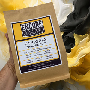 Hand holding a 12 oz bag of Encore Coffee & Chocolate Ethiopia Shakiso Guji coffee in front of a flowing geometric background with swirls of yellow, black, and cream.