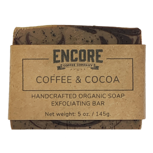 A bar of handcrafted soap wrapped in a kraft label with an Encore logo on the top. The label states "coffee & cocoa" below the logo, followed by a description of the soap as handcrafted and its weight. The soap bar is rectangular with smooth edges and a textured surface, and it appears to be made with natural ingredients. The kraft label adds a rustic touch to the overall appearance of the soap.