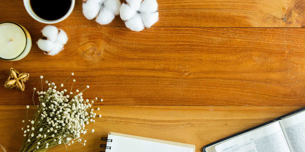 Flat lay of a cozy workspace with a cup of black coffee, cotton flowers, an open Bible, candle, dried flowers, and a notebook on a wooden table.
