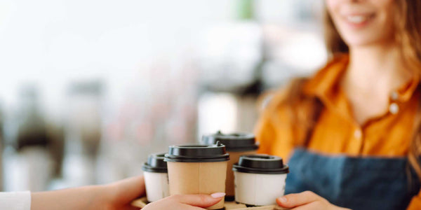 Barista handing a tray of takeaway coffee cups to a customer in a warm, inviting café setting.