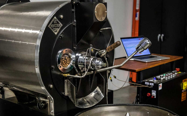 Commercial coffee roaster from US Roaster Corp in operation with green coffee beans inside, monitored by a connected laptop for roast profiling.