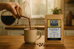 Hand pouring black coffee from a Chemex into a ceramic mug beside a labeled Encore Coffee & Chocolate Guatemala Lake Atitlán bag on a wooden kitchen counter.