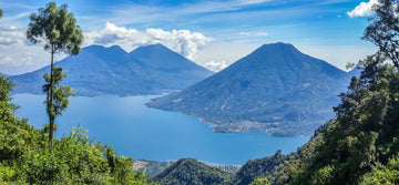 Scenic view of a large blue lake surrounded by forested mountains and volcanoes under a partly cloudy sky.