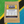 Encore Coffee & Chocolate’s Tanzania coffee bag on a vibrant painted background of green, yellow, black, and blue inspired by the Tanzanian flag. Label details include origin (Mbozi, Songwe), flavor notes (apricot, cocoa, brown sugar, black tea), and roast date 