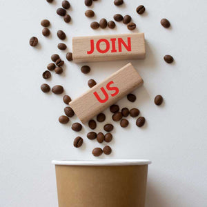 Coffee beans falling into a paper cup with wooden blocks reading 'JOIN US' – coffee recruitment or community concept
