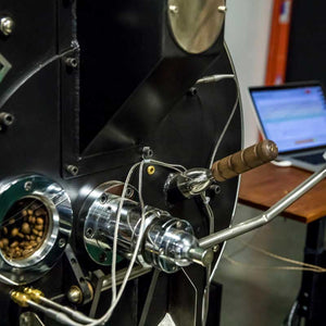 Close-up of a commercial coffee roasting machine with green coffee beans inside and a laptop monitoring roast data in the background