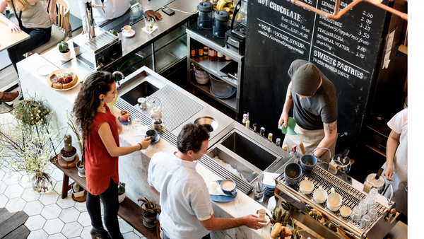 Overhead view of a busy specialty coffee shop with baristas, customers, espresso equipment, pastries, and a hot chocolate menu.