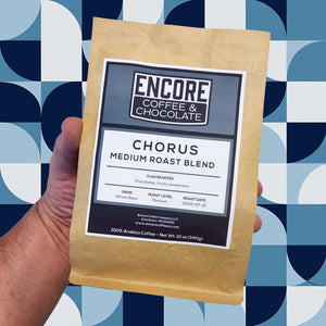 Hand holding a bag of Encore Coffee & Chocolate Chorus Medium Roast Blend against a blue geometric background.
