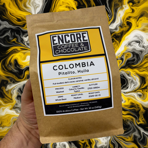 Hand holding a brown kraft bag of Encore Coffee & Chocolate Colombia Pitalito, Huila whole bean coffee against a yellow, black, and white swirling background.