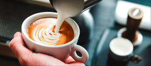 A close-up of a barista pouring steamed milk from a metal pitcher into a white cup of coffee with latte art, set in a cafe environment.