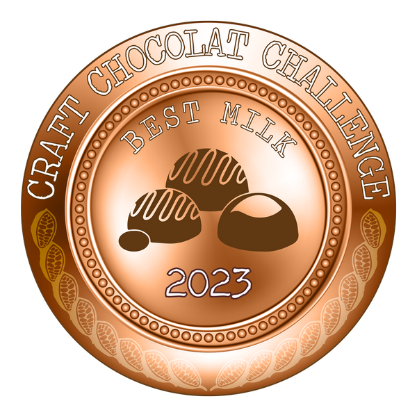 An image of a bronze medal with the text 'Best Milk Chocolate 2023 Craft Chocolat Challenge' engraved on it. The medal features a design with bonbons in the middle, surrounded by a ring of cacao plants. The medal is sitting on top of a bronze-colored milk chocolate bar with a glossy finish. The chocolate bar is sitting on a white background with no other visible objects in the image. The brand name or logo is not visible, and the product name is not provided in the image.