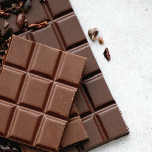 A close-up photograph of a chocolate bar with a textured surface, surrounded by scattered cocoa nibs and dusted with cocoa powder. The chocolate bar is broken into several uneven pieces, revealing a smooth and glossy interior. The lighting is soft and warm, casting a gentle shadow on the surface beneath the chocolate.